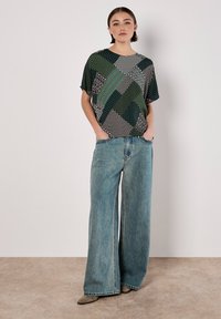 Abstraktes gemustertes Oberteil in Grün, Schwarz und Weiß; lockere Passform. Kombiniert mit weit geschnittenen, hellblauen Jeans mit rohem Saum.