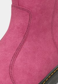 Pink suede ankle boot with a textured finish, featuring black stitching and a rubber sole with yellow accents. Curved shape at the top.