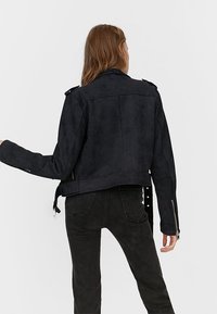 Giacca corta in stile scamosciato nera con spalline, zip asimmetrica e una finitura leggermente testurizzata. Presenta maniche lunghe e una vestibilità rilassata.