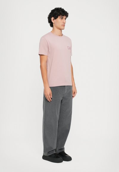 Lys rosa bomull T-skjorte med korte ermer og en logo foran, sammen med brede grå kordfløyelsbukser og svarte sneakers.