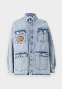 Gaiši zila pārāk liela džinsū jacket ar apkakli, priekšējo pogu aizdari, četriem kabatām un brūnu ādas plāksteri kreisajā krūtī.