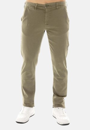 Uomo che indossa pantaloni verde oliva aderenti e sneakers bianche, in piedi davanti a uno sfondo chiaro e uniforme.