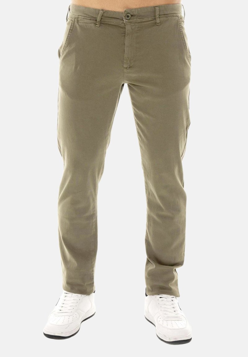 Homme portant un pantalon ajusté vert olive et des baskets blanches, debout devant un fond clair uni.