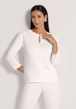 Vrouw met lang donker haar, gekleed in een wit shirt met lange mouwen en een kleine keyhole, en een witte broek, poseert tegen een effen lichte achtergrond.