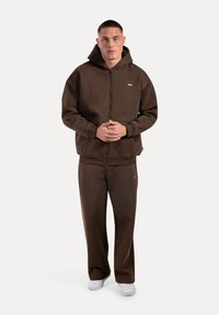 Ein junger Mann steht mit Blick nach vorne, trägt einen braunen Zip-Hoodie und passende Hosen mit weißen Turnschuhen, die Hände vor sich gefaltet.