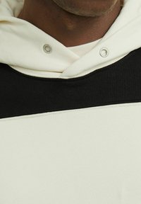 Hoodie noir et crème avec un tissu texturé. Il présente une capuche avec un cordon crème et des œillets métalliques près de l'encolure.