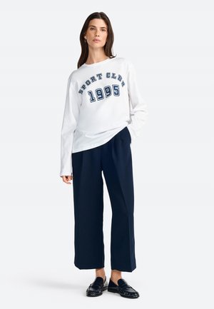 White long-sleeve shirt with "SPORT CLUB 1995" print in dark blue, paired with navy wide-leg trousers and black loafers.