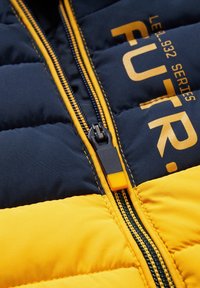 Gros plan sur la fermeture éclair d'une veste matelassée noire et jaune, avec le texte "FUTR" et des coutures jaunes sur le tissu.