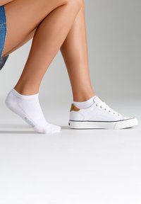 Calzino bianco alla caviglia con logo di lato, abbinato a sneaker in tela bianca con un accento beige sul tallone e rifiniture nere.