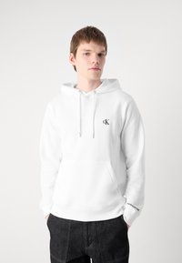 Sudadera blanca con capucha y cordón, bolsillo frontal y pequeño logo negro de CK. Cuenta con puños y dobladillo acanalados, fabricada en un suave tejido texturizado.