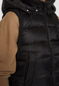 Gilet imbottito nero con finitura lucida, chiusura con zip, bottoni a pressione e cappuccio. Presenta fasce regolabili e tasche laterali.