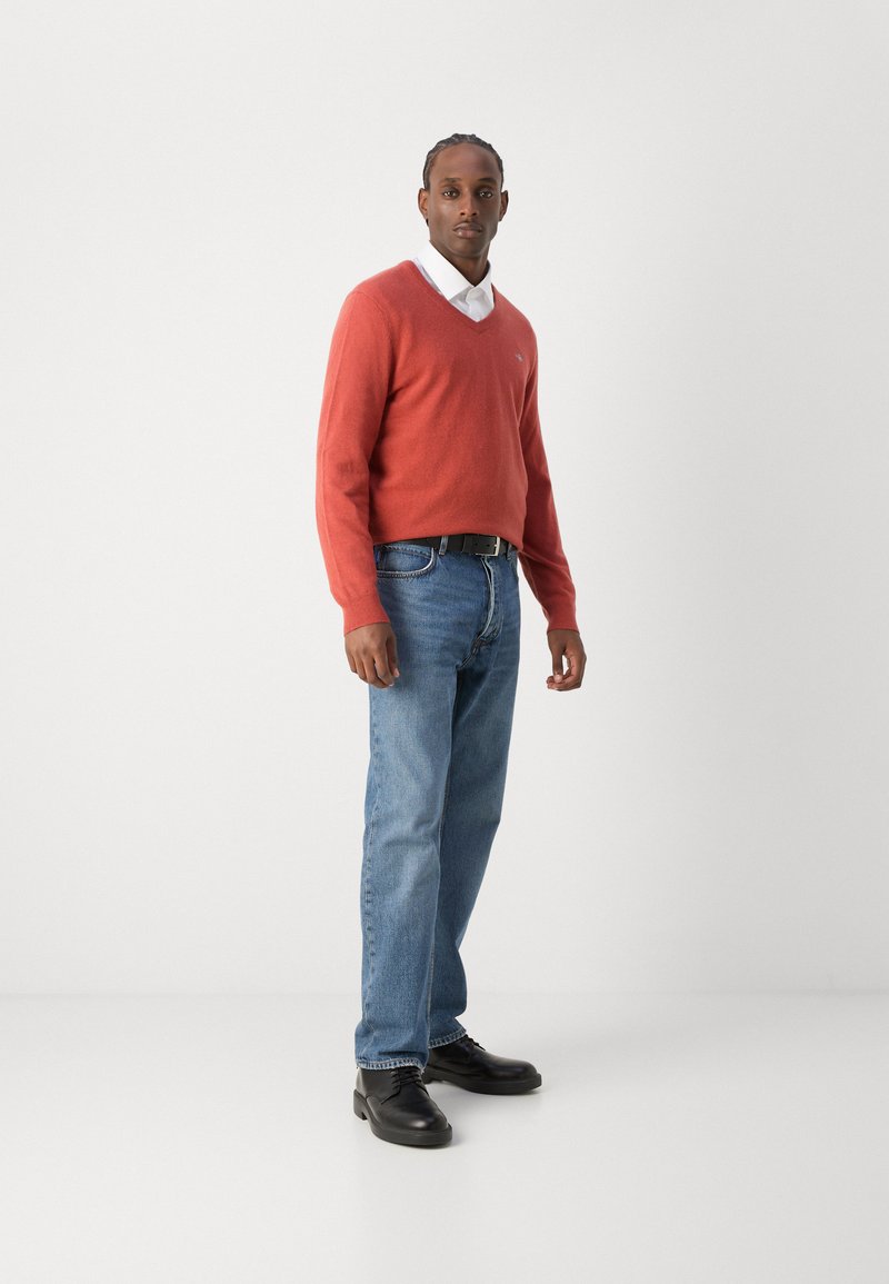 Roter Pullover mit V-Ausschnitt, getragen über einem weißen Hemd. Blaue Jeans und schwarze Schuhe. Einfaches Design ohne auffällige Muster oder Akzente.