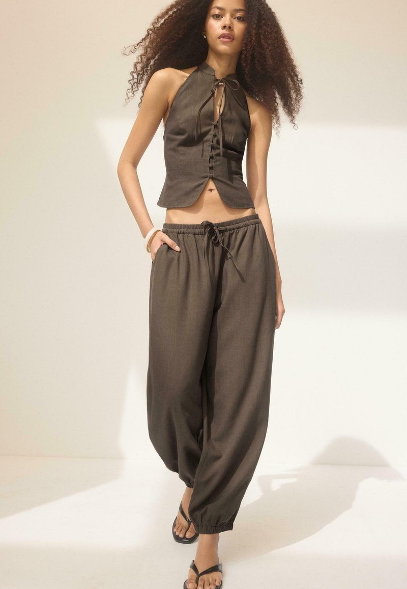 Jeune femme aux cheveux bouclés portant un haut sans manches marron foncé à col halter et un pantalon ample, marchant en avant avec une main dans la poche.