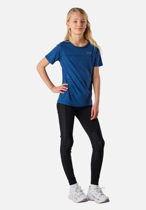 Adolescente debout, la main sur la hanche, portant un t-shirt bleu à manches courtes, des leggings noirs et des chaussures de sport blanches sur fond blanc.