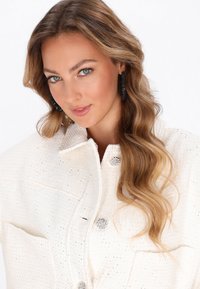 Witte textuurjack met glanzende embellissments en zilveren knopen; model met lang golvend haar en grote zwarte oorbellen.