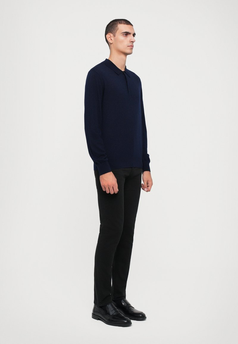 Polo a maniche lunghe blu navy in tessuto morbido, abbinato a pantaloni neri aderenti e scarpe di pelle nere, in piedi su uno sfondo bianco.