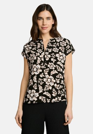 Femme aux cheveux bruns mi-longs portant un chemisier noir à manches courtes avec de grands motifs floraux blancs et un pantalon noir.