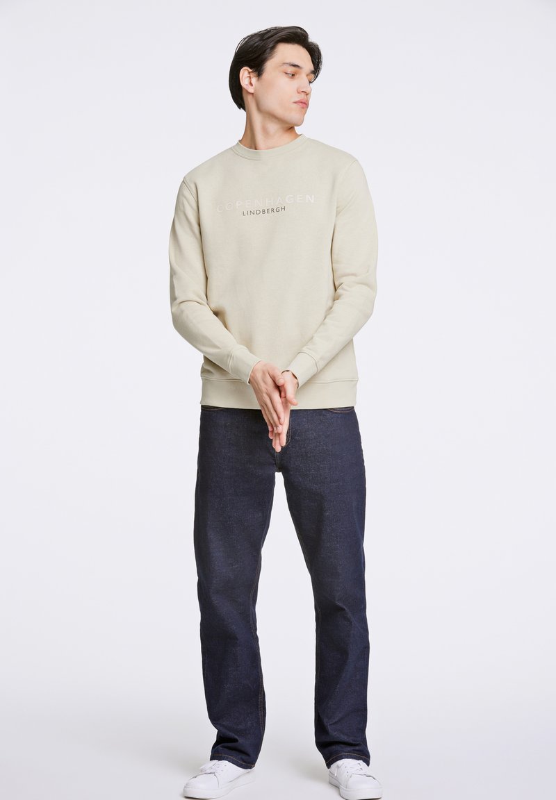 Felpa beige con scollatura rotonda, con la scritta "Copenhagen" e "Lindbergh" stampata. Abbinata a jeans blu scuro e scarpe da ginnastica bianche.