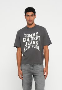 Šedé bavlnené tričko s krátkymi rukávmi a okrúhlym výstrihom, na ktorom je výrazný biely nápis: "TOMMY ATH.DEPT JEANS NEW YORK." Nosené so svetlošedými džínsami.