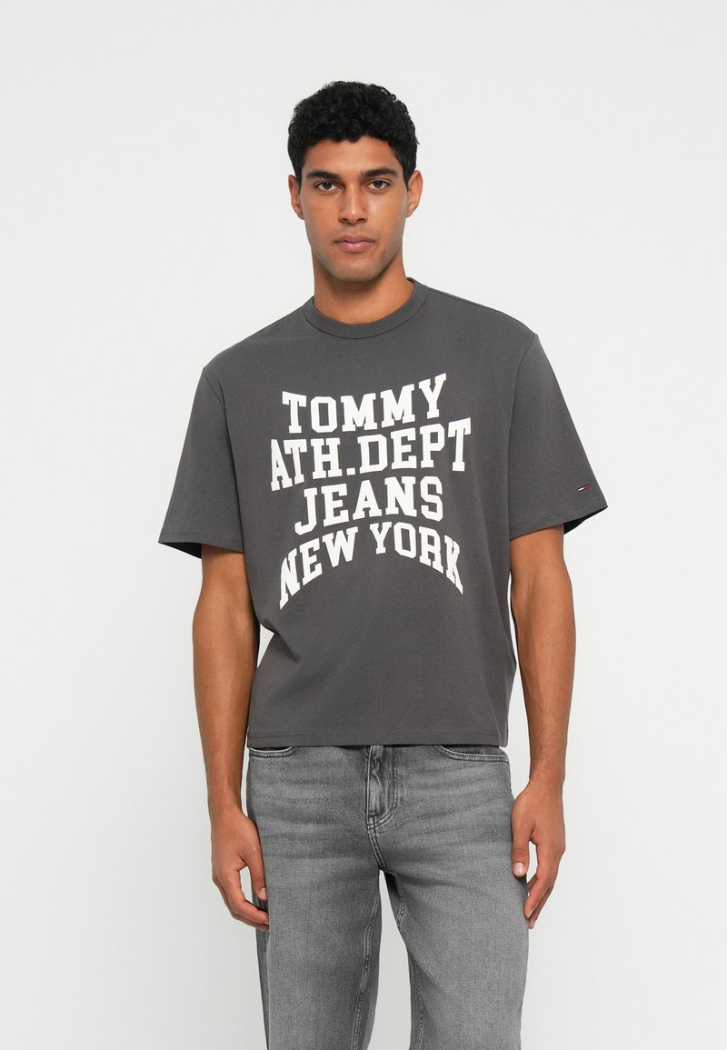 Šedé bavlnené tričko s krátkymi rukávmi a okrúhlym výstrihom, na ktorom je výrazný biely nápis: "TOMMY ATH.DEPT JEANS NEW YORK." Nosené so svetlošedými džínsami.
