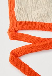 Kreemivärvi kootud kangas paksu oranži äärisega. Omab pikkasid oranže nööre, tasast tekstuuri ja kontrastset värviskeemi.