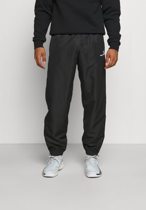 Persona con pantaloni da jogging neri e scarpe da ginnastica bianche, in piedi contro uno sfondo chiaro e semplice, con le mani rilassate lungo i fianchi.