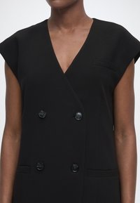 Μαύρο αμάνικο διπλό γιλέκο με τέσσερα κουμπιά και V-λαιμόκοψη, που φοριέται από ένα άτομο με μέτριο προς σκουρόχρωμο τόνο δέρματος.