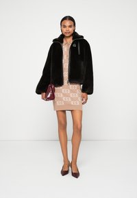 Black faux fur bomber jacket with a zip closure and hood, worn over a patterned tan dress. Accessorized with a small burgundy handbag.