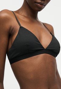 Κοντινό πλάνο ενός ατόμου που φοράει μαύρο σουτιέν τριγωνικό με λεπτές τιράντες πάνω σε μονόχρωμο φόντο.