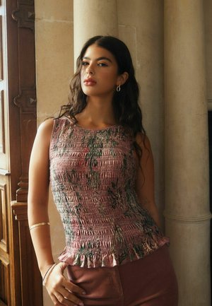 Young woman wearing a sleeveless pink and green textured top, standing indoors near a wooden door and stone columns.
