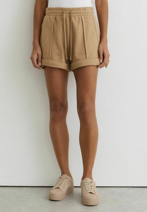 Person trägt beige Shorts mit umgekrempeltem Saum und Kordelzugbund sowie beige Sneakers, steht vor einer weißen Wand auf grauem Boden.