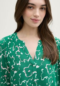 Grüne Bluse mit floralem Muster in Weiß und Pink, V-Ausschnitt, Knopfverschluss und weichem, strukturiertem Stoff.