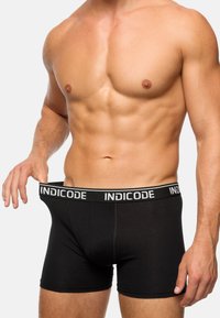 Boxers negros hechos de una tela suave, con una cinturilla blanca en contraste que tiene impreso "INDICODE". Largo hasta el muslo y ajuste ceñido.
