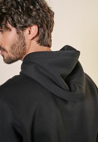Svart hoodie med en mjuk textur, som har en stor huva och en enkel design. Tyget verkar tjockt och varmt, lämpligt för avslappnad klädsel.