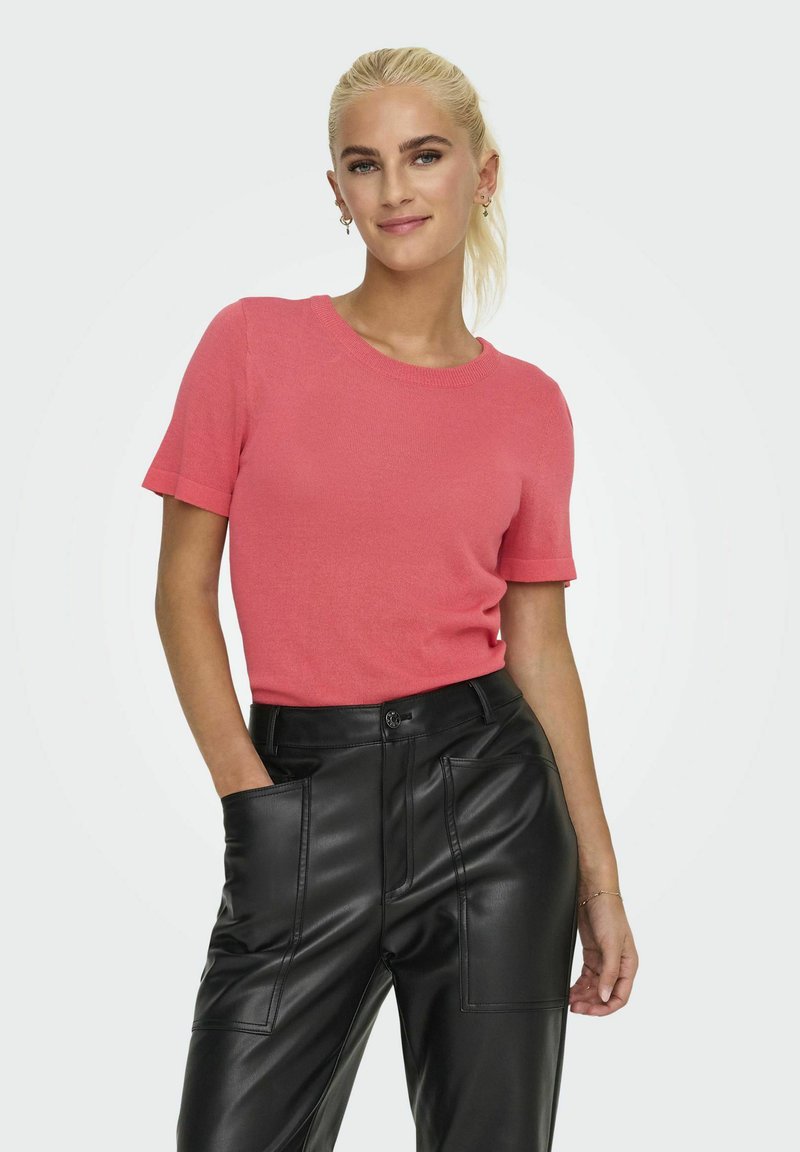 Top de punto coral de manga corta con cuello redondo, combinado con pantalones de cuero negros que tienen bolsillos delanteros y un ajuste relajado.