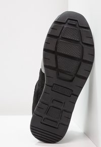 Podeszwa czarnego buta z głębokimi bieżnikami oraz wytłoczonym logo "GUESS" na środku, na tle białego tła.