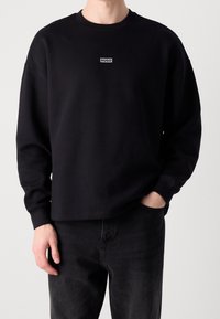 Hombre con un sudadera negra de mangas largas con el logo "HUGO" en el pecho y jeans negros, con una mano en el bolsillo, frente a un fondo sencillo.