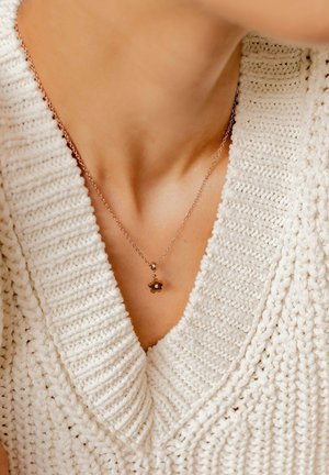 Rosenvergoldete Blumenkette mit einem kleinen Edelstein, die auf einem strukturierten, cremefarbenen Strickpullover mit V-Ausschnitt liegt.