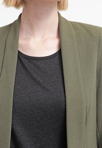 Olivově zelený blazer vyrobený z lehkého materiálu s hladkou texturou, nošený přes černou košili, s otevřeným předním dílem a bez viditelných zavírání.