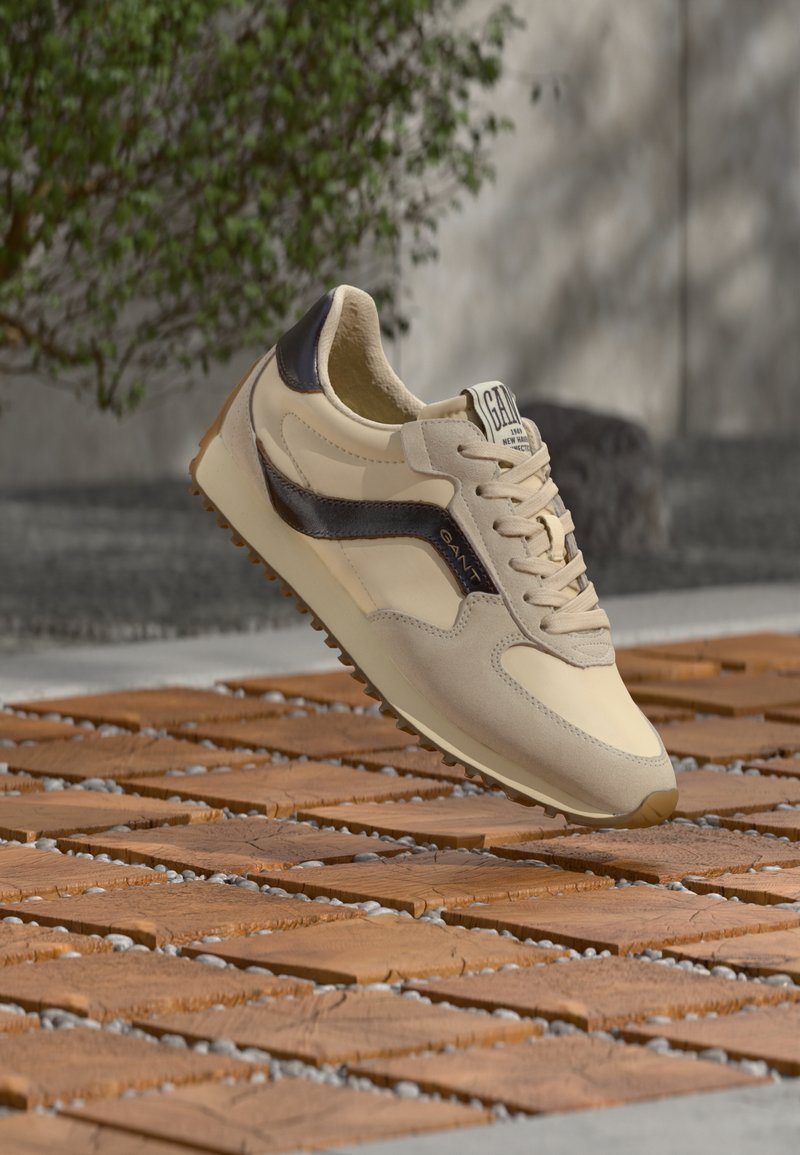 Chaussure de sport beige avec des accents marron foncé, design à lacets et semelle texturée, reposant sur une surface en bois avec des cailloux.