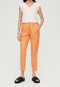 Witte v-hals top met oranje strepen, gecombineerd met oranje linnen broek met een elastische tailleband en zijzakken, gedragen met zwarte sandalen.