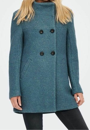 Femme portant un manteau en laine bleu sarcelle à double boutonnage avec des boutons noirs, debout, les mains le long du corps, sur fond blanc uni.
