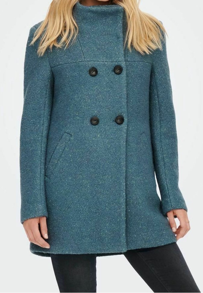 Femme portant un manteau en laine bleu sarcelle à double boutonnage avec des boutons noirs, debout, les mains le long du corps, sur fond blanc uni.