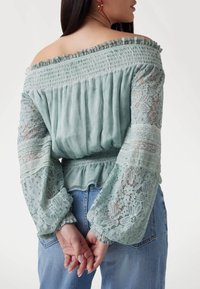 Light green off-the-shoulder blouse with smocked upper, layered lace long sleeves, and a fitted waist, paired with blue jeans.