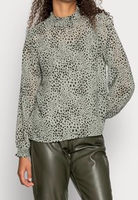 Blusa transparente de color verde claro con estampado de animal en negro, cuello alto y mangas largas con fruncidos. Combinada con pantalones de cuero verde oscuro.