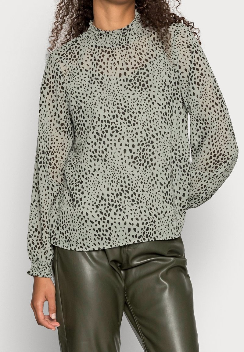 Blusa transparente de color verde claro con estampado de animal en negro, cuello alto y mangas largas con fruncidos. Combinada con pantalones de cuero verde oscuro.
