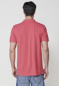 Moška polo majica s kratkimi rokavi v koralno rožnati barvi iz bombaža, z običajnim ovratnikom in nekoliko prilega silhueto. Vidne podrobnosti šivanja.