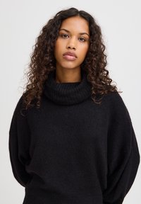Pull en tricot noir avec un col roulé côtelé haut. Présente des manches larges et une coupe décontractée, mettant en valeur un matériau doux et texturé.