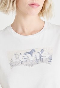Balta medvilninė marškinėliai su mėlynu grafiniu piešiniu, kuriame pavaizduoti du arkliai ir augalai, su „Levi's“ prekės ženklo pavadinimu ryškiomis raidėmis.