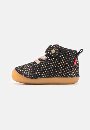 Zapato negro con patrón de estrellas, con parte superior de tela suave, cordones beige, tira de Velcro y suela de goma. Acentuado con una etiqueta de marca roja.